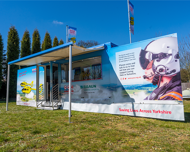 Photo shows one of the Yorkshire Air Ambulance promotional vehicles parked on grass. The sides are out and display photos of the yellow helicopter and a pilot with a helmet on and has some wording, which cannot be made out on the photo. The words Saving Lives Across Yorkshire are along the bottom. There is an awning out from the side of the vehicle. There are open doors with steps and a large window. On top of the vehicle are two flags. 