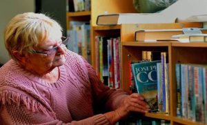 A lady with short light hair, wearing a pink jumper places a book on to a shelf containing other books.