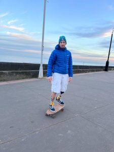 A man wearing a blue puffa jacket, blue woolly hat, white or grey shorts, yellow socks and dark trainers is riding a skateboard and has a prosthetic leg.