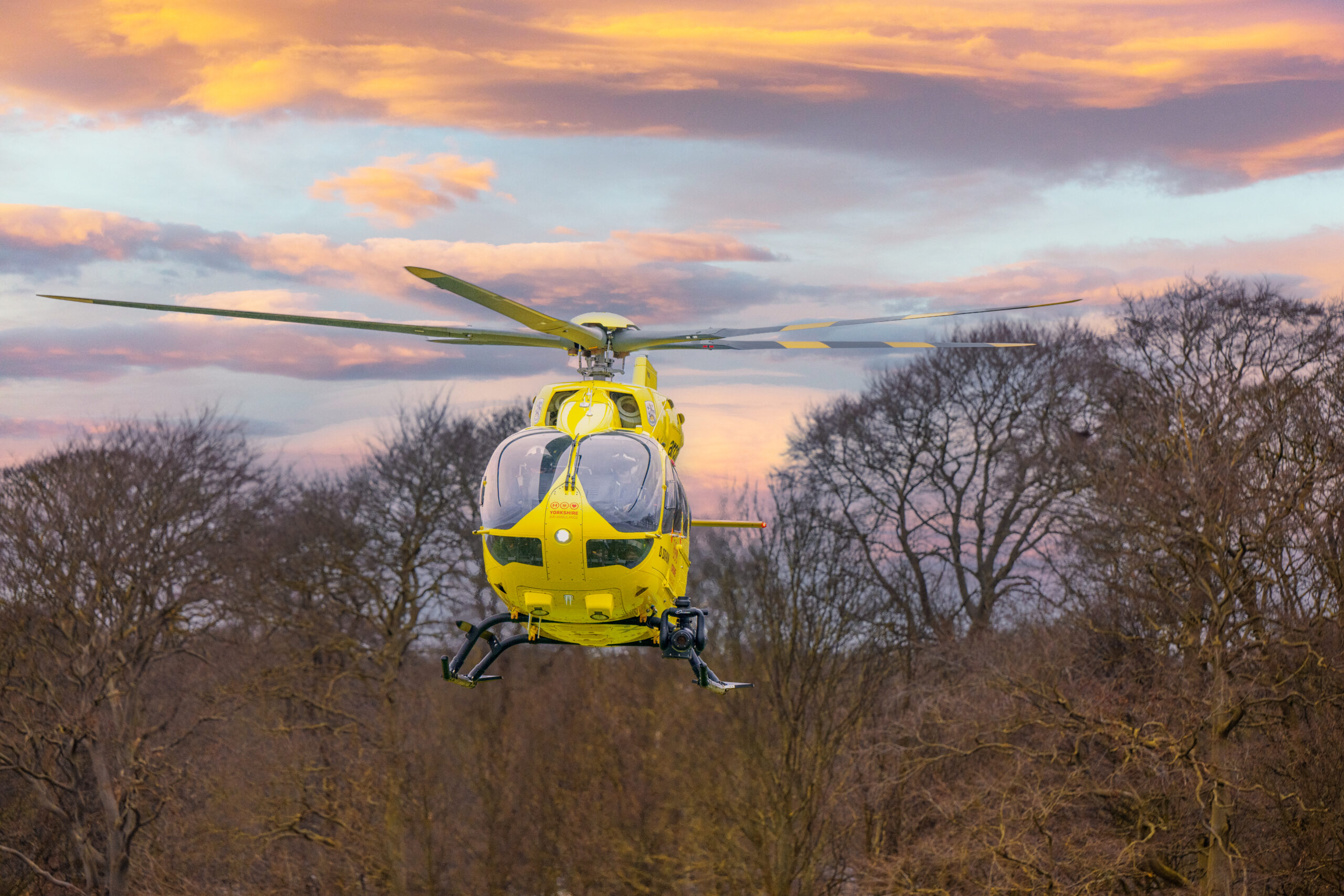 A front on image of a yellow helicopter flying, with trees in the background.