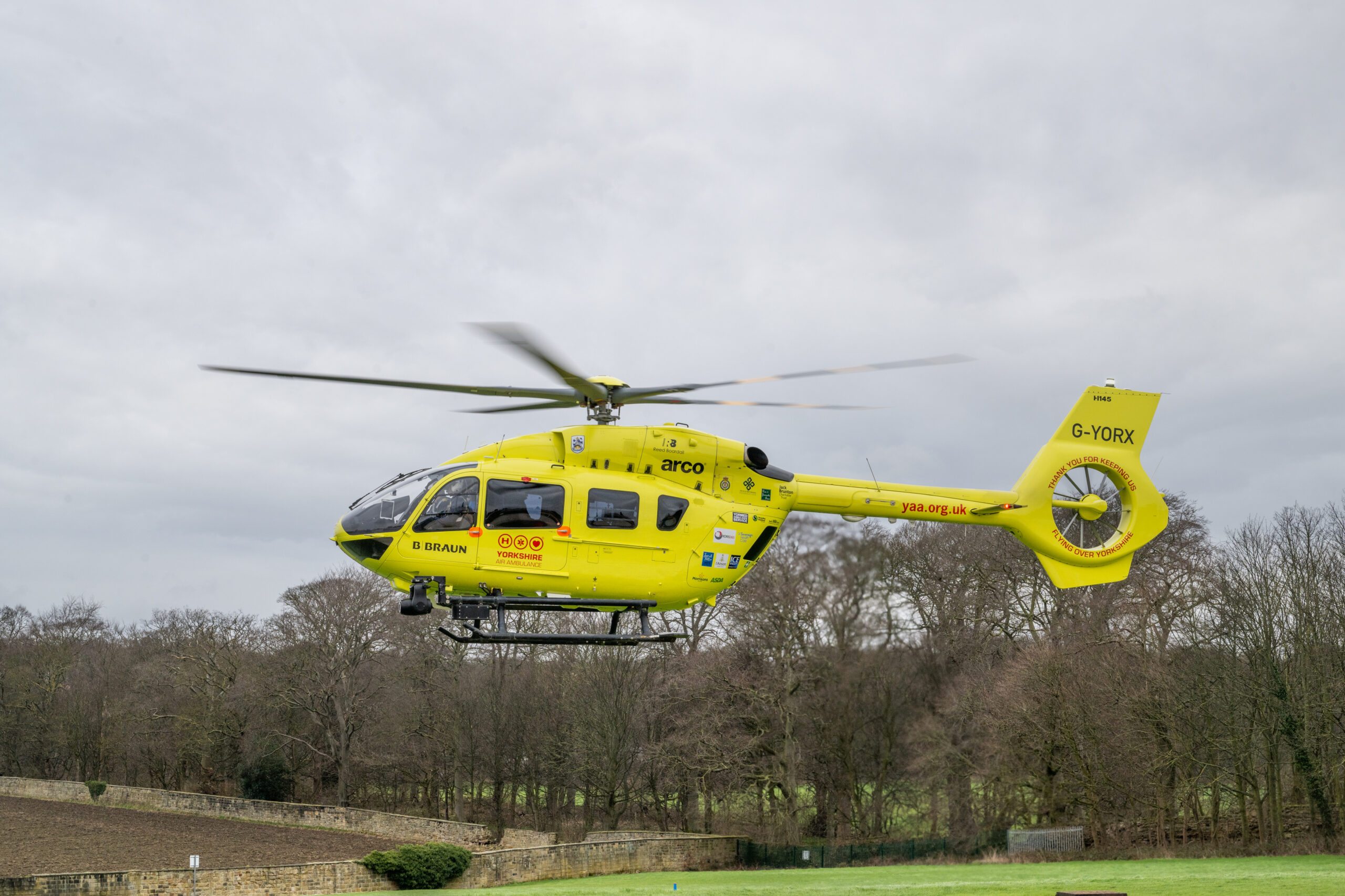 A yellow helicopter is flying above a field with trees in the background.