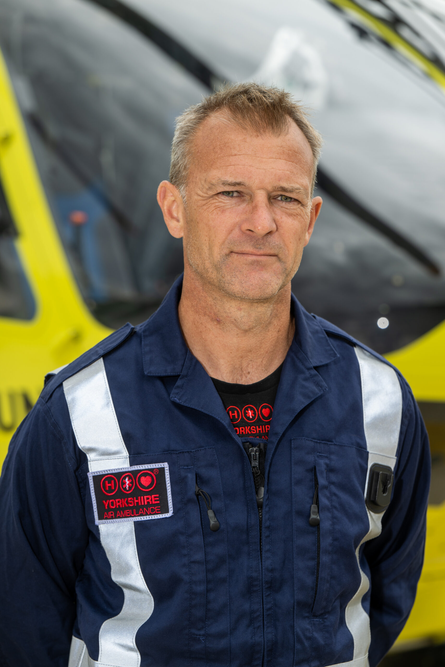 A man with short hair, wearing a blue flying suit, is standing in front of a yellow helicopter