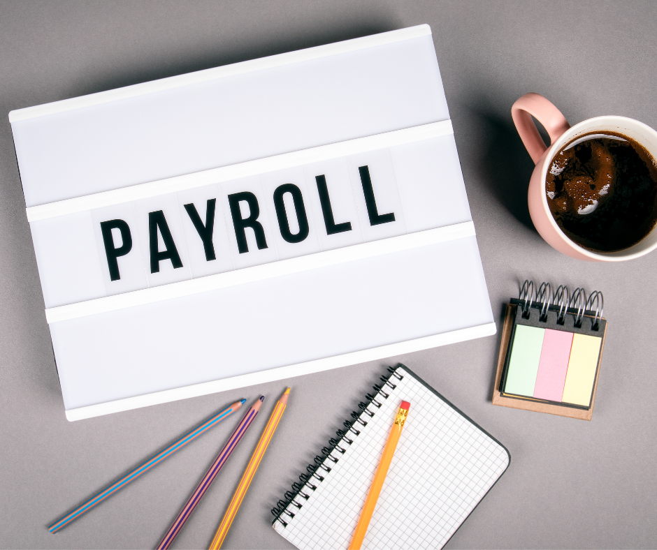 Overhead shot of a lightbox with the word PAYROLL on it, next to a cup of coffee and notebook and some pencils, all laid on a grey table top.