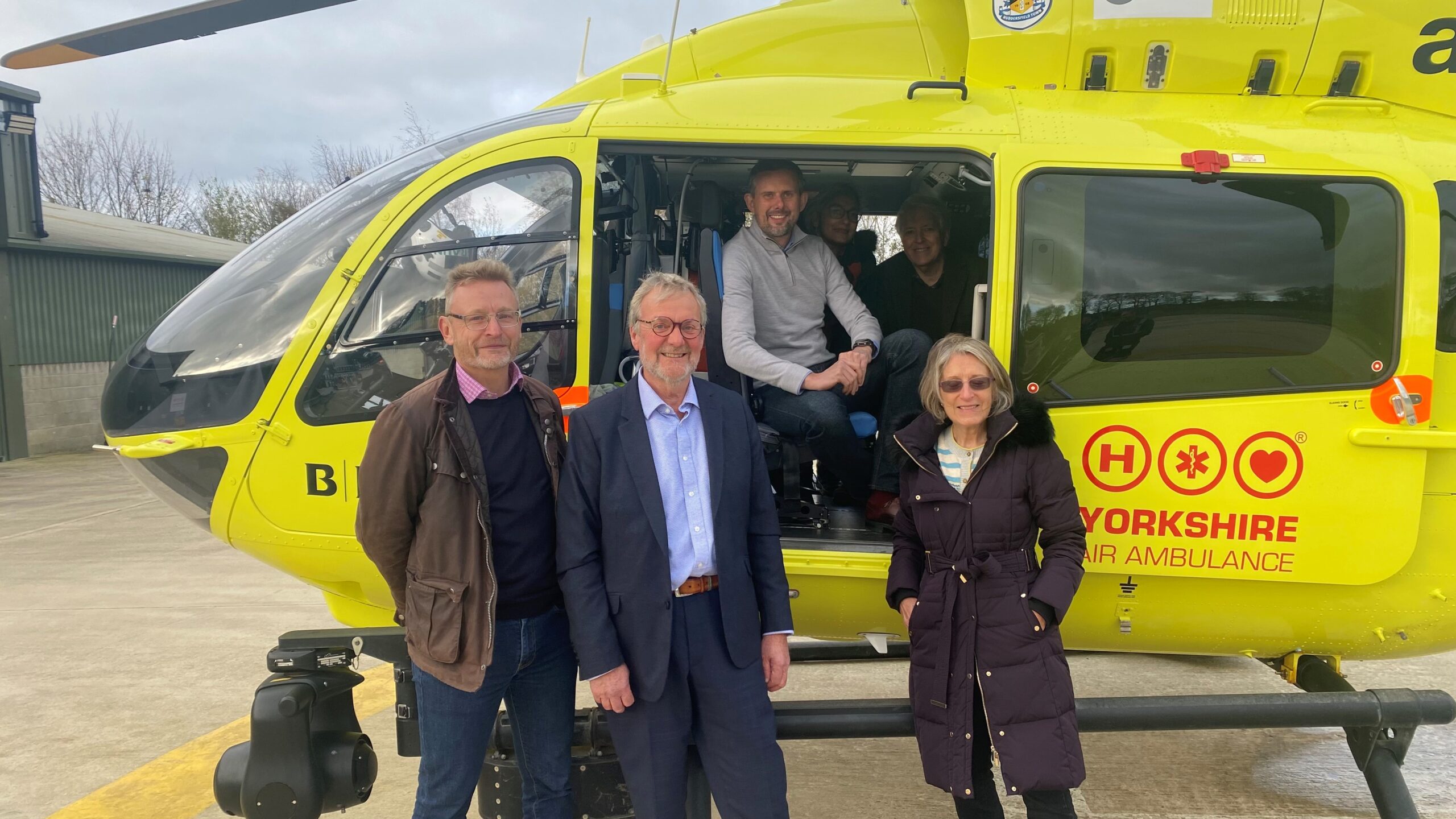 A group of people are stood in front of a yellow helicopter and some are sat in the aircraft, with the door open.