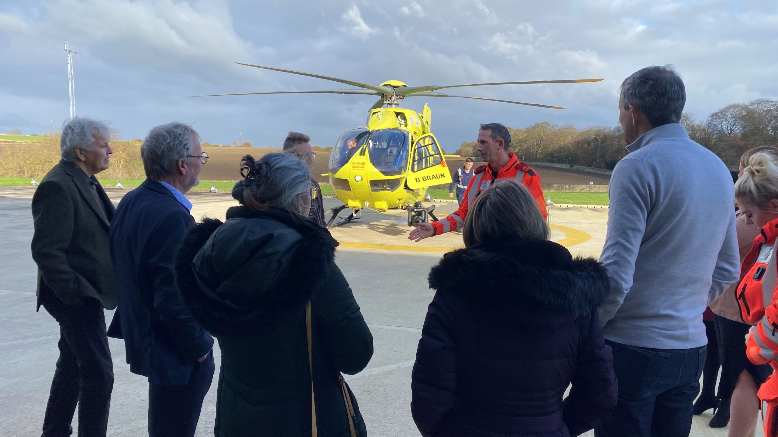 A group of people are listening to a man in an orange flight suit giving a talk in front of a yellow helicopter. 