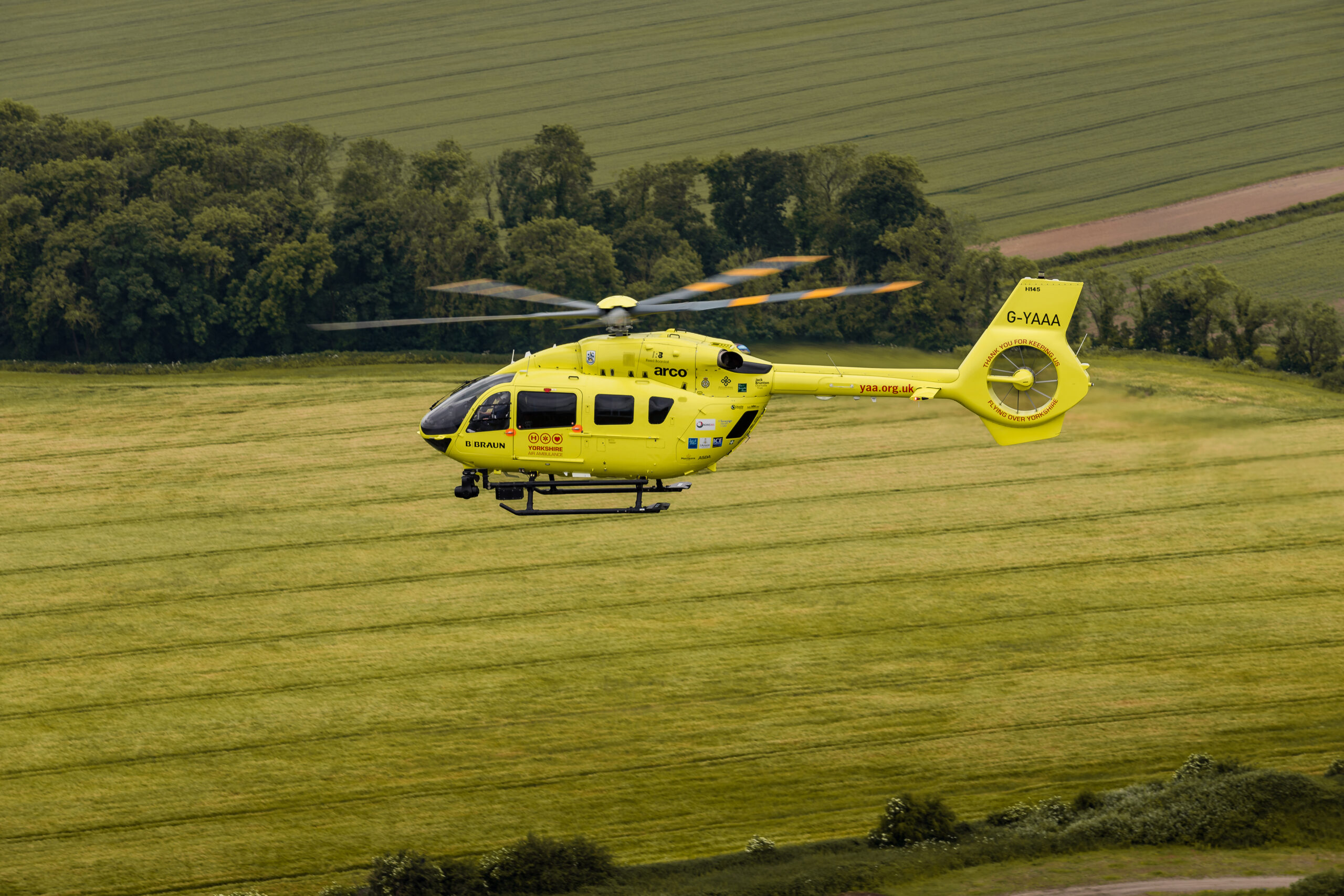 A yellow helicopter is hovering above a field surrounding by trees