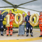 A group of people are standing in front of a yellow helicopter, holding gold balloons. There are a '1' and two '0' balloons. Some of the people are wearing flight suits.