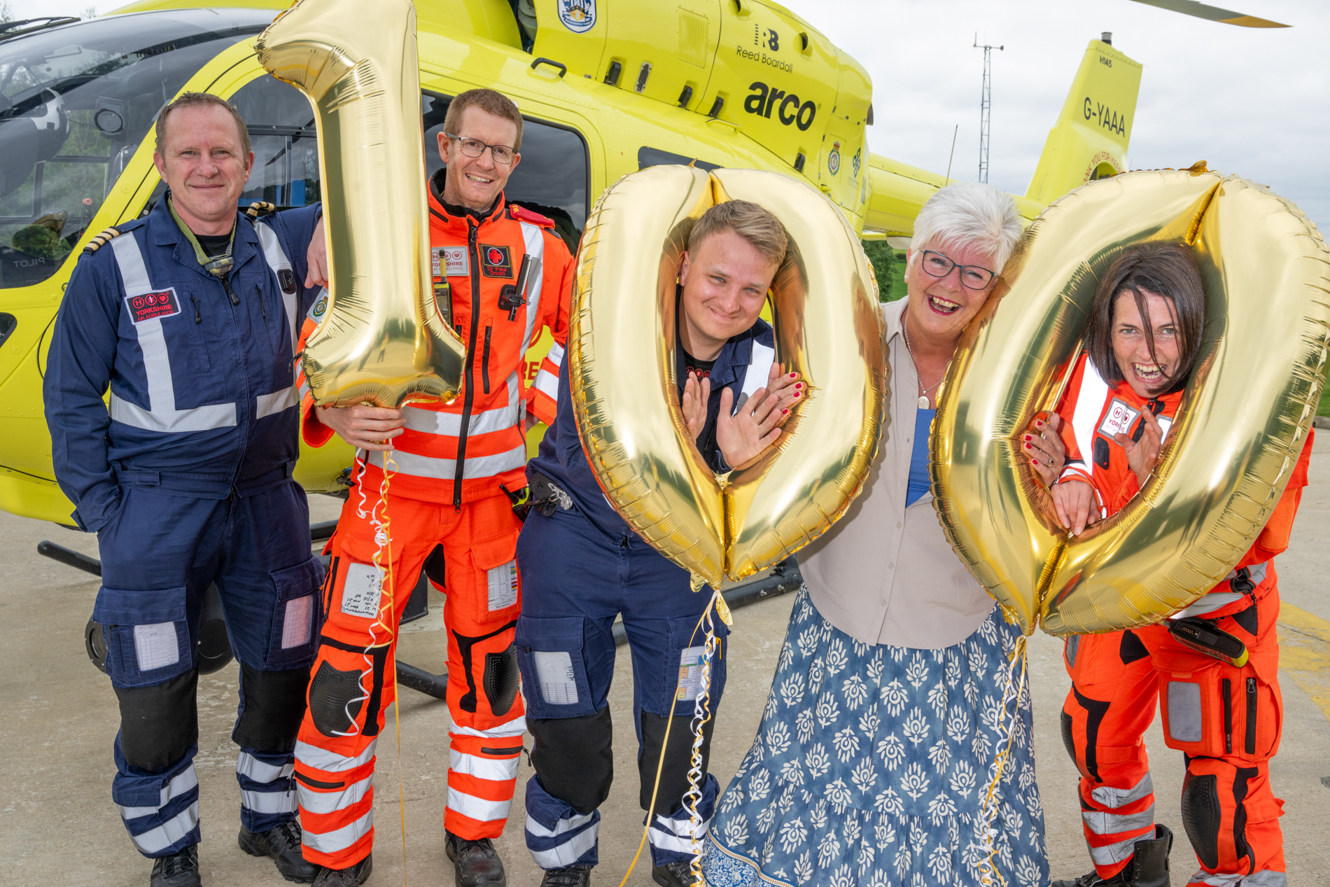 A group of people are standing in front of a yellow helicopter, holding gold balloons. There are a '1' and two '0' balloons. Some of the people are wearing flight suits. two of them have their heads through the two '0' balloons.