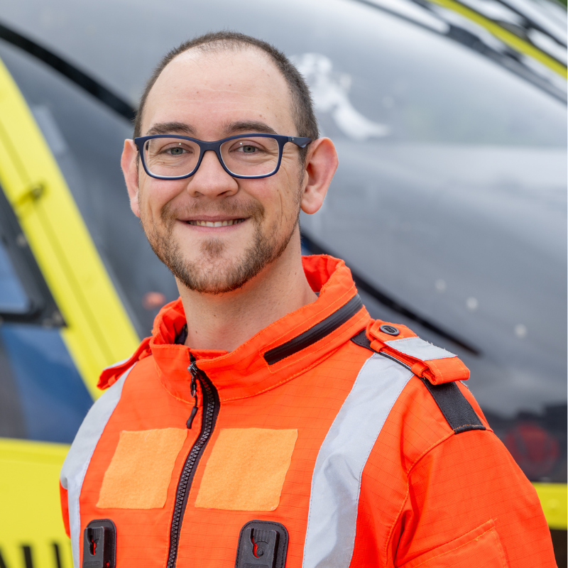 A man wearing glasses and an orange flight suit is standing in front of a yellow helicopter