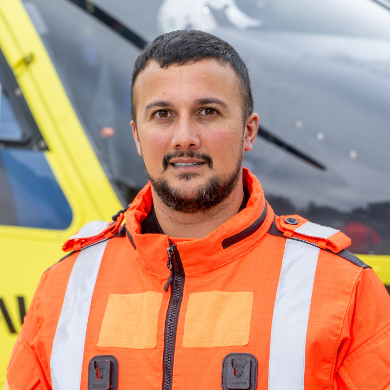 A man with dark hair, beard and moustache, wearing an orange flight suit is standing in front of a yellow helicopter