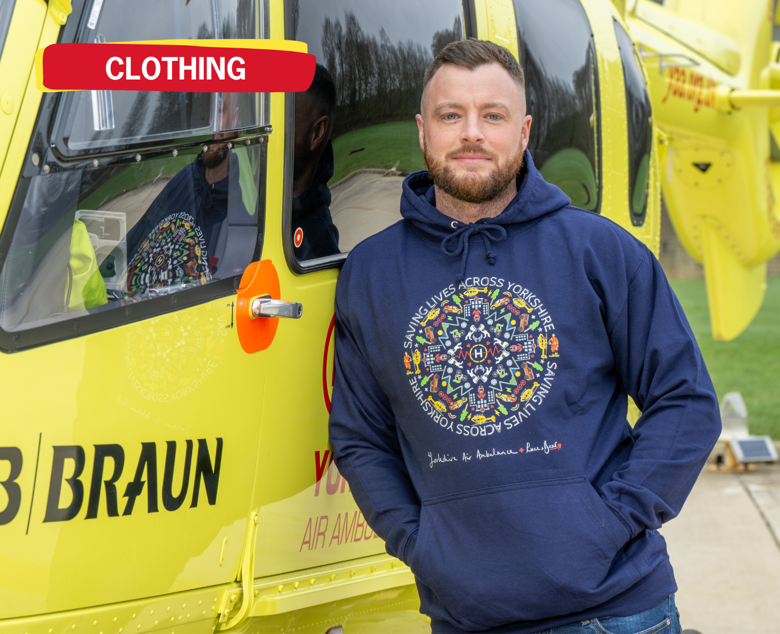 A man wearing a blue hoodie with a pattern on the front is leaning against a yellow helicopter. There is a graphic in the top left hand corner with the word 'CLOTHING'