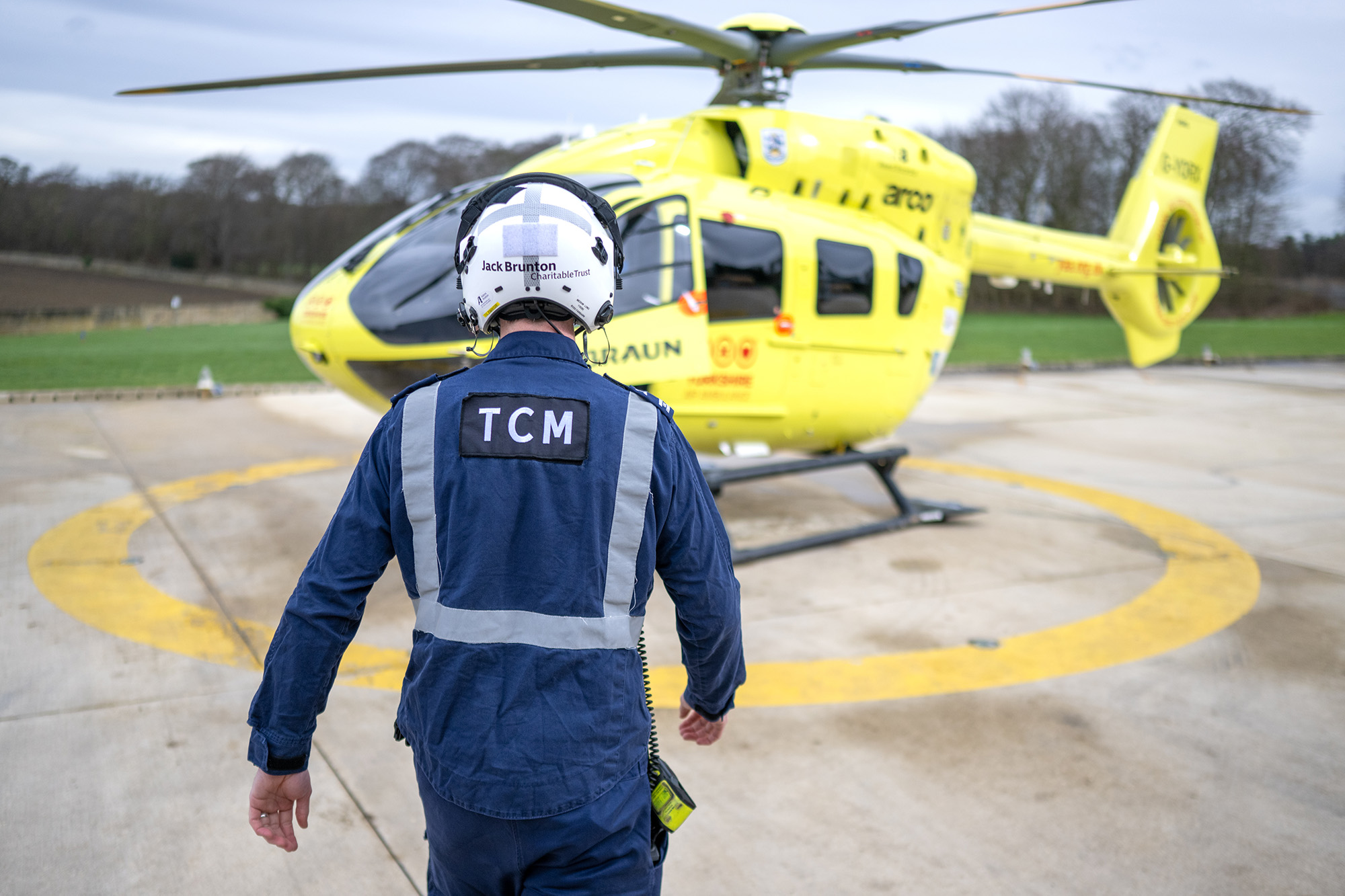 A person wearing a flight helmet and a blue flight suit with the letters 'TCM' on the back is walking towards a yellow helicopter.