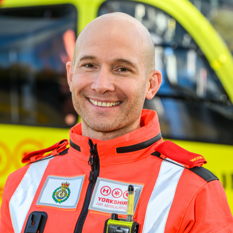 A man in an orange doctors flight suit stood in front of a yellow helicopter