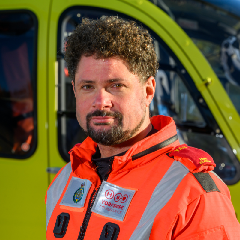 A man in an orange doctors flight suit stood in front of a yellow helicopter