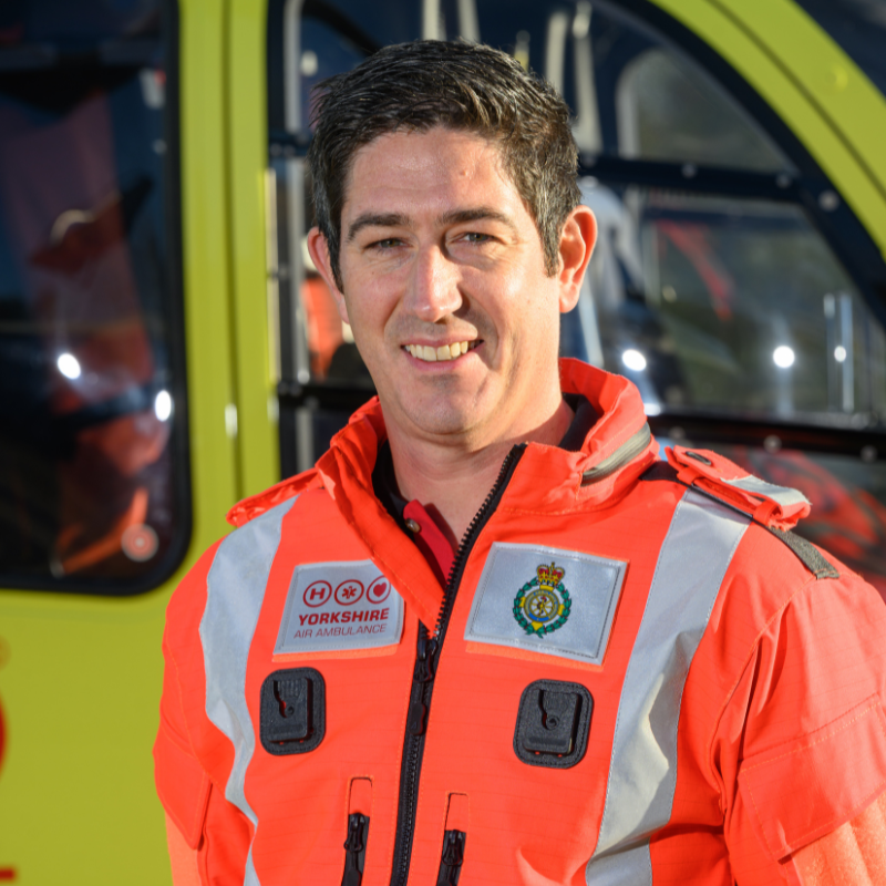 A man in an orange doctors flight suit stood in front of a yellow helicopter