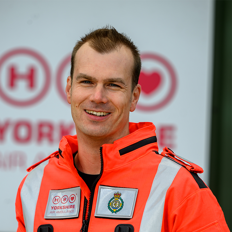 A man with short dark hair, wearing an orange flight suits which has an ambulance service badge on.