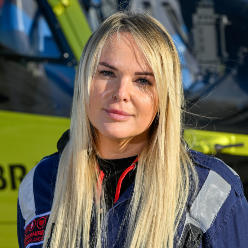 Head and shoulders image of a lady with long blonde hair wearing a dark blue flight suit infront of a yellow helicopter