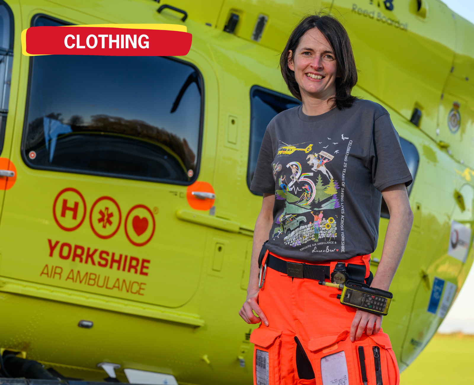 A lady with dark hair wearing orange flight trousers and a grey t-shirt with a design featuring a yellow helicopter, is standing in front of a yellow helicopter